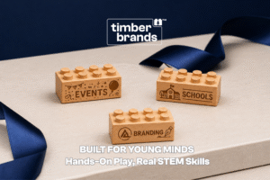 Three Timber Brands wooden building bricks engraved with "Events," "Schools," and "Branding" icons on a neutral surface with navy ribbon, beneath the words "Built for Young Minds."