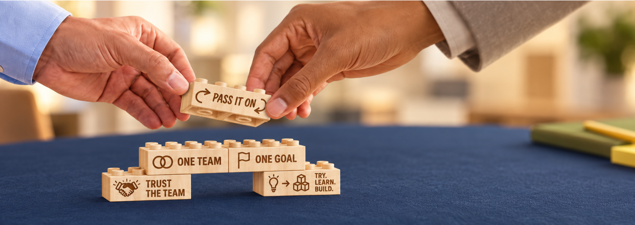 Hands passing a Pass It On wooden brick above connected bricks engraved with One Team, One Goal, Trust the Team, and Try. Learn. Build.