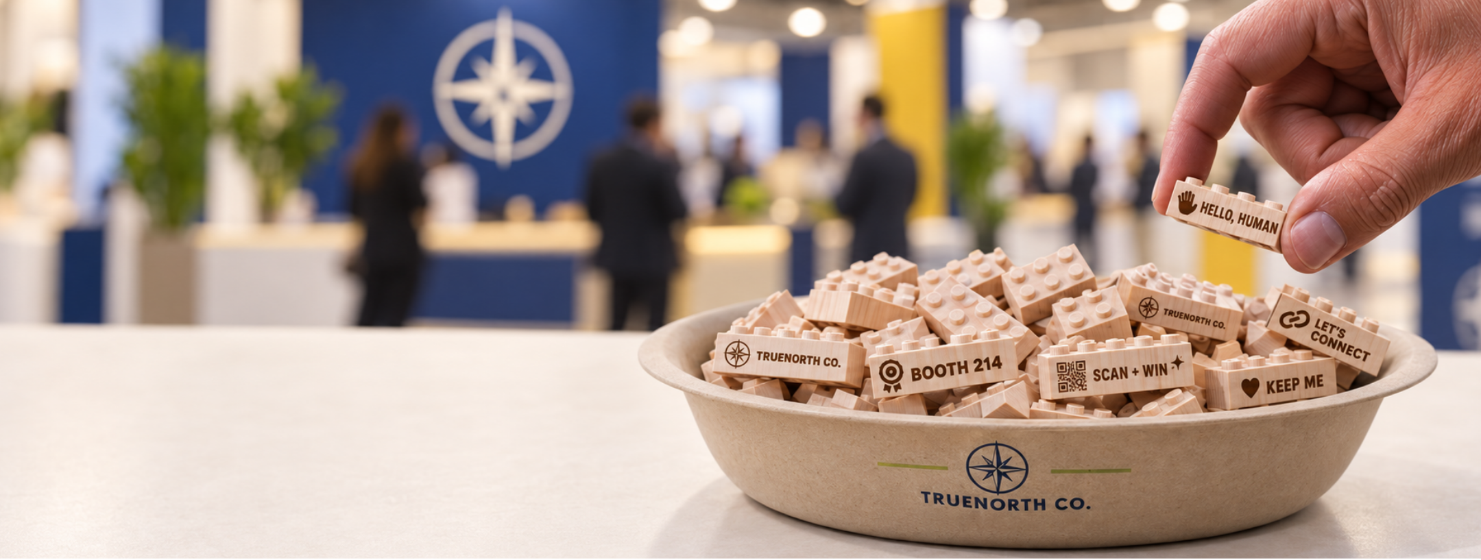 A hand selecting an engraved wooden brick giveaway from a bowl on a trade show booth counter.