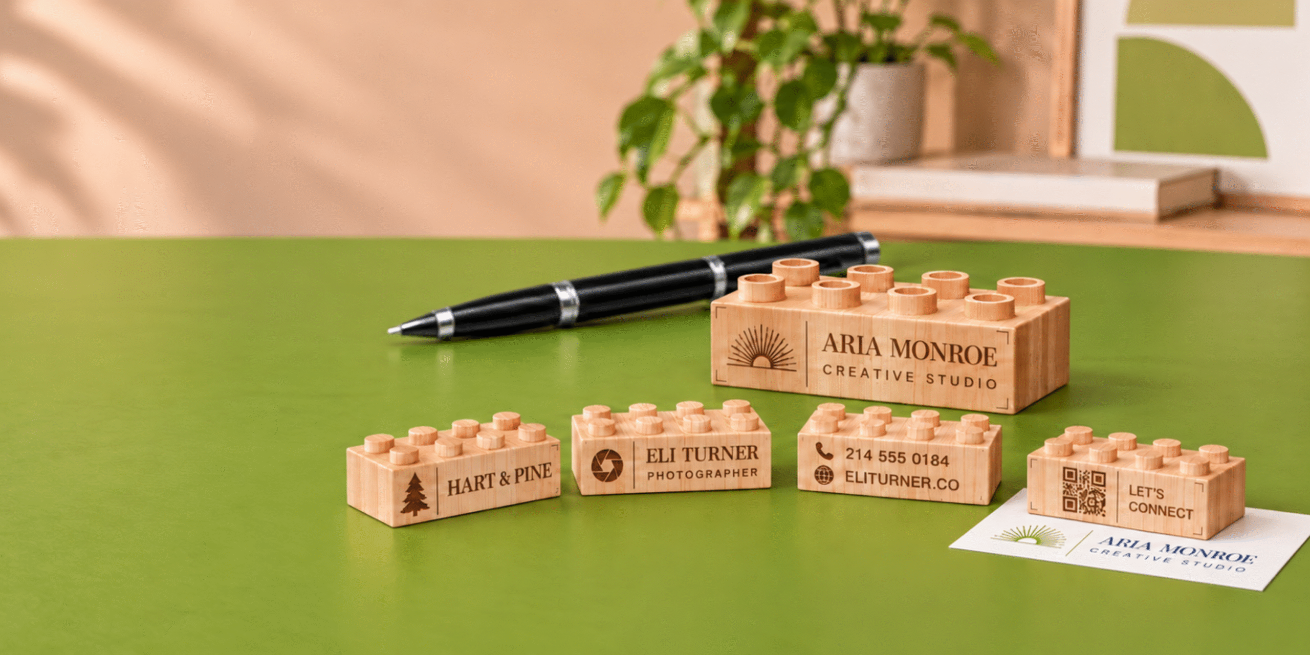 Wooden brick business cards with engraved names, logos, contact details, and a QR-code design on a green desk.